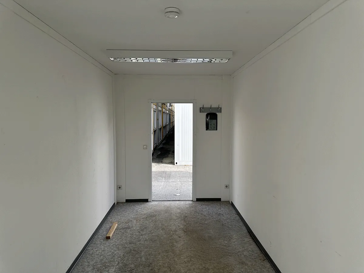 Innenansicht eines grauen Bürocontainers mit Blick auf die geschlossene weiße Eingangstür und die Deckenbeleuchtung.