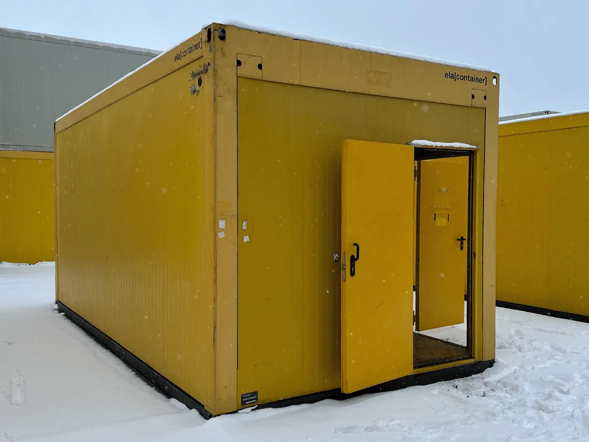 Rückansicht des gelben 20-Fuß-Bürocontainers mit geschlossener weißer Eingangstür auf einem Betriebsgelände.