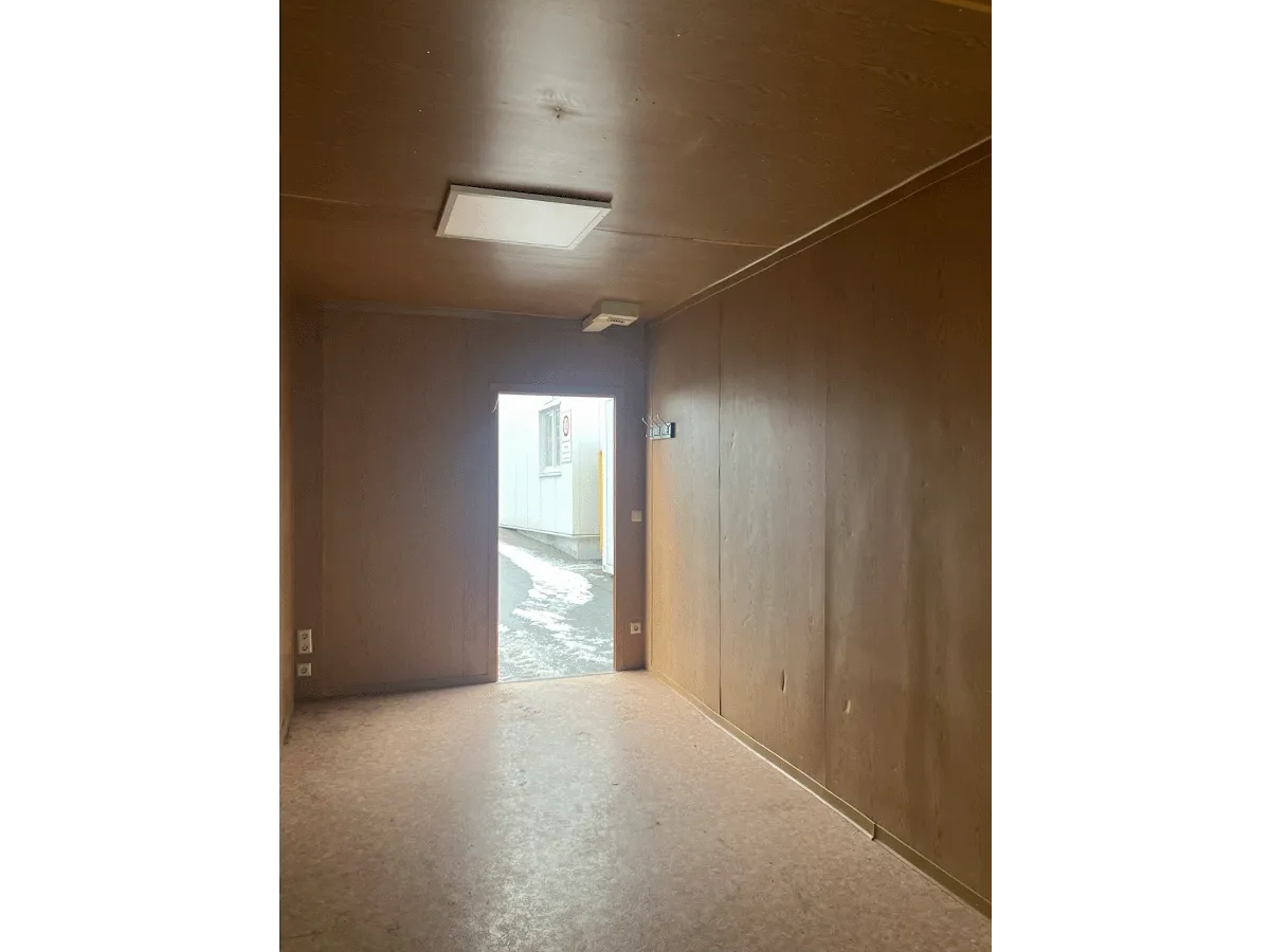 Innenansicht eines grauen Bürocontainers mit Blick auf die geschlossene weiße Eingangstür und Deckenleuchte.
