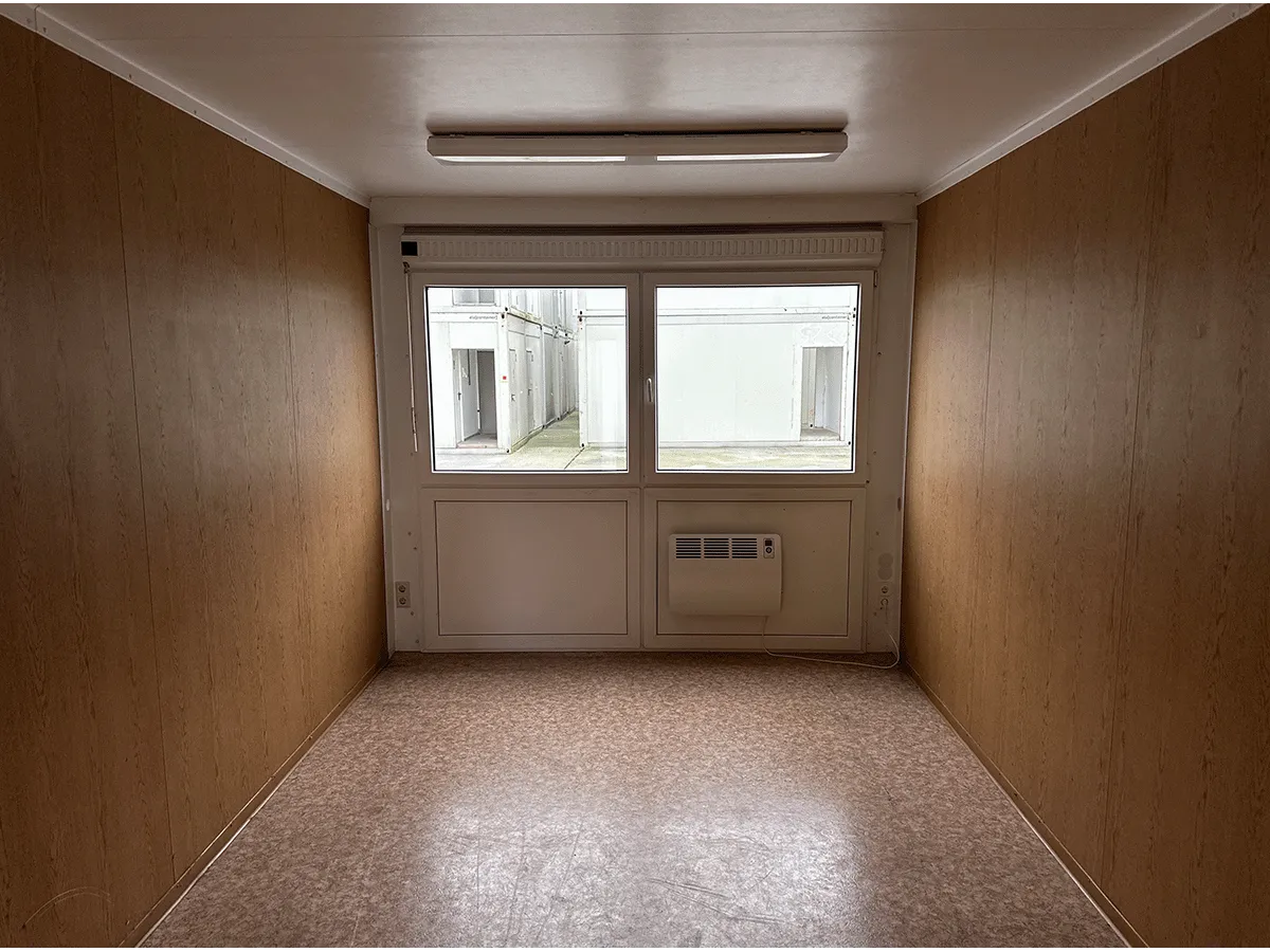 Innenansicht eines leeren grauen Bürocontainers mit Fenster, Heizung und hellem Bodenbelag.