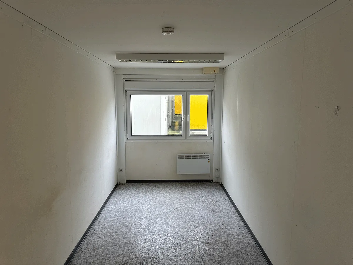 Innenansicht eines grauen Bürocontainers mit Blick auf die geschlossene weiße Eingangstür und Deckenleuchte.