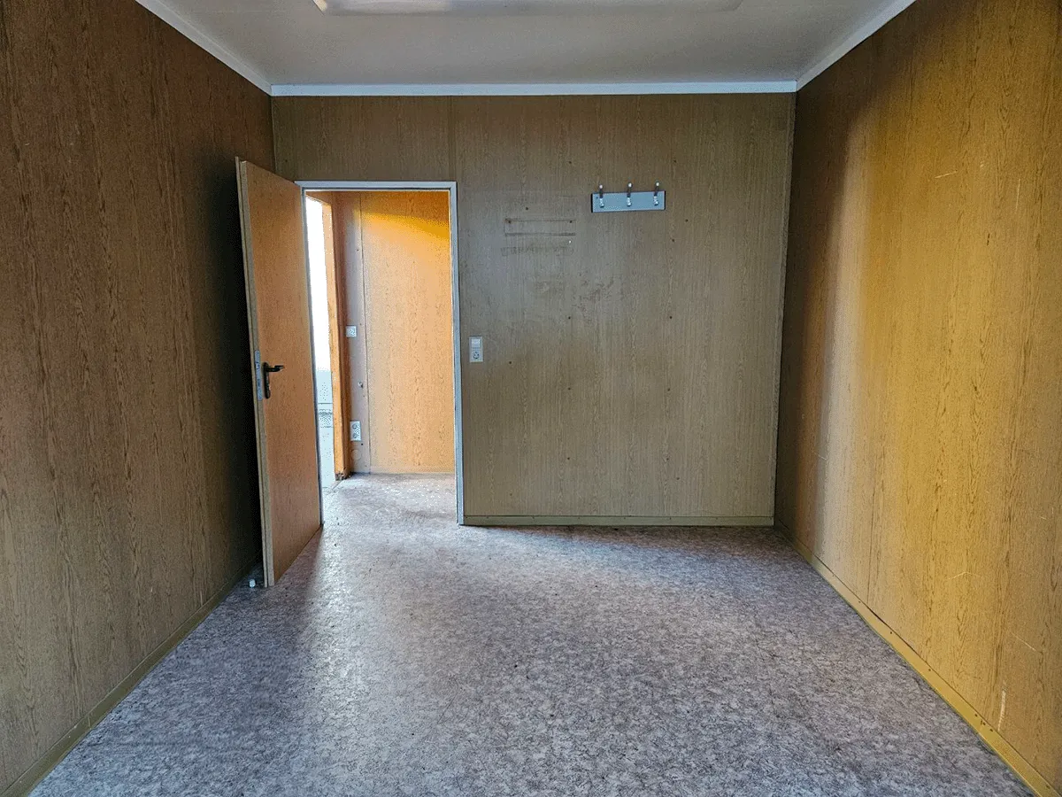 Innenansicht des gelben 20-Fuß-Bürocontainers (3m Breite) mit Blick auf die geschlossene Eingangstür und den Sicherungskasten.