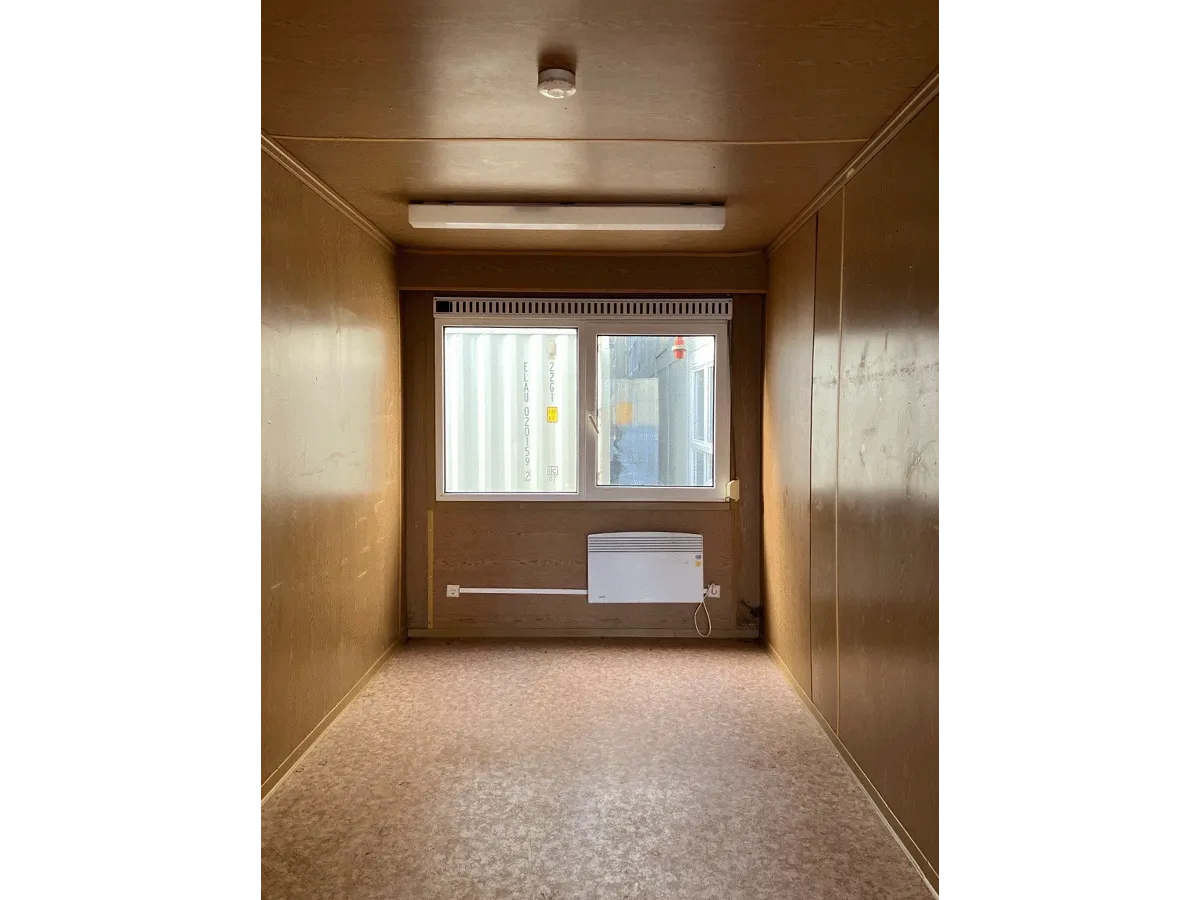 Innenansicht eines leeren gelben Bürocontainers mit Fenster, Heizung und hellem Bodenbelag.