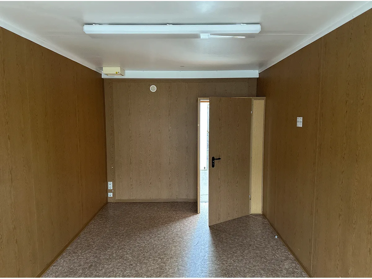 Innenansicht eines grauen Bürocontainers mit Blick auf die geschlossene Eingangstür und die Deckenbeleuchtung.