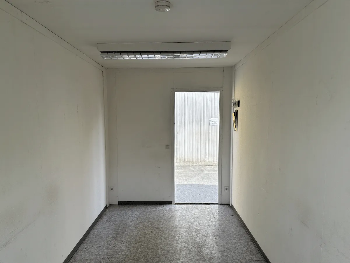 Innenansicht eines leeren grauen Bürocontainers mit Blick auf die geöffnete Tür und Deckenleuchte.