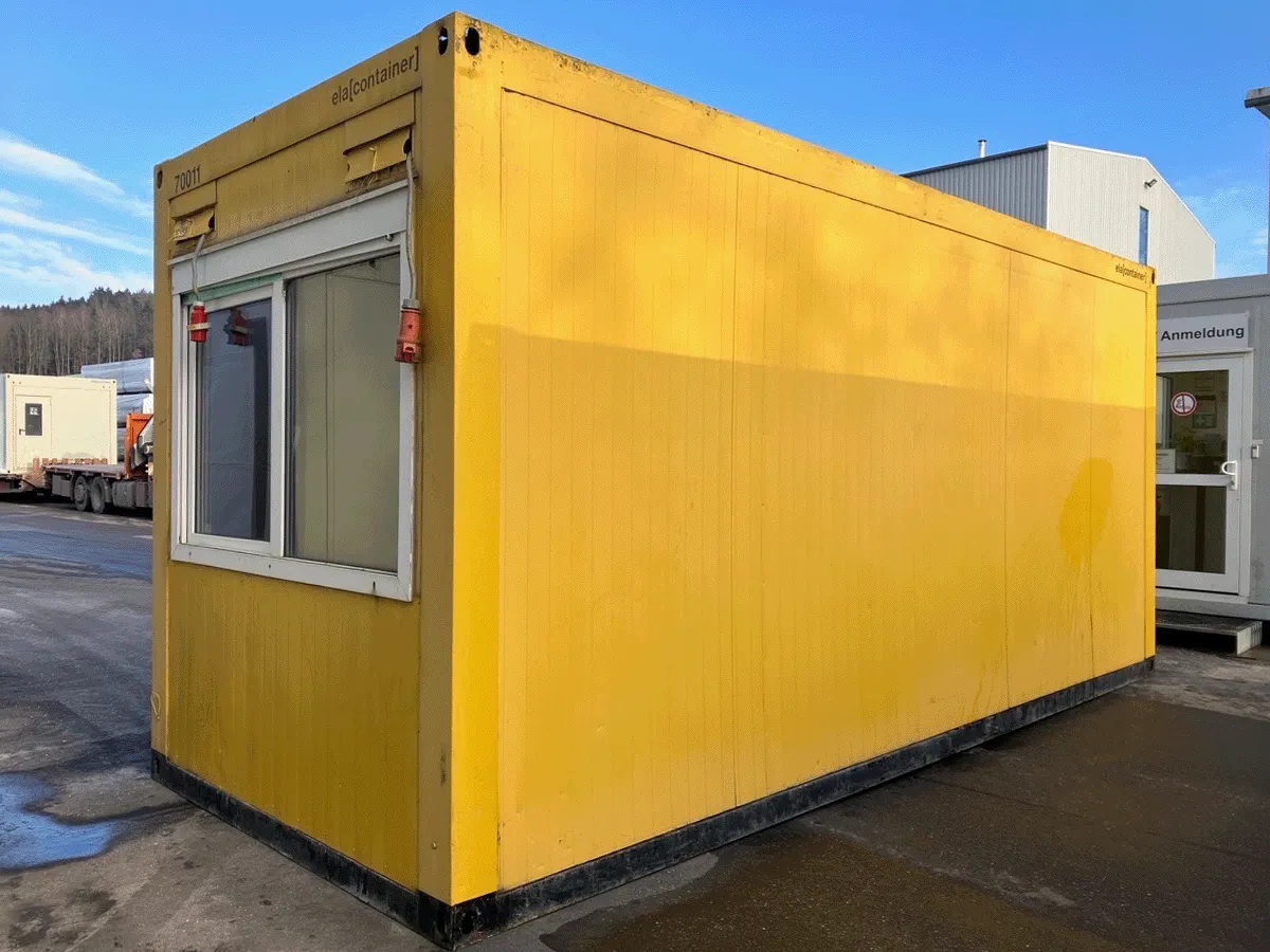 Außenansicht eines gebrauchten gelben Bürocontainers mit Fenster und Rollladen auf einem asphaltierten Gelände.