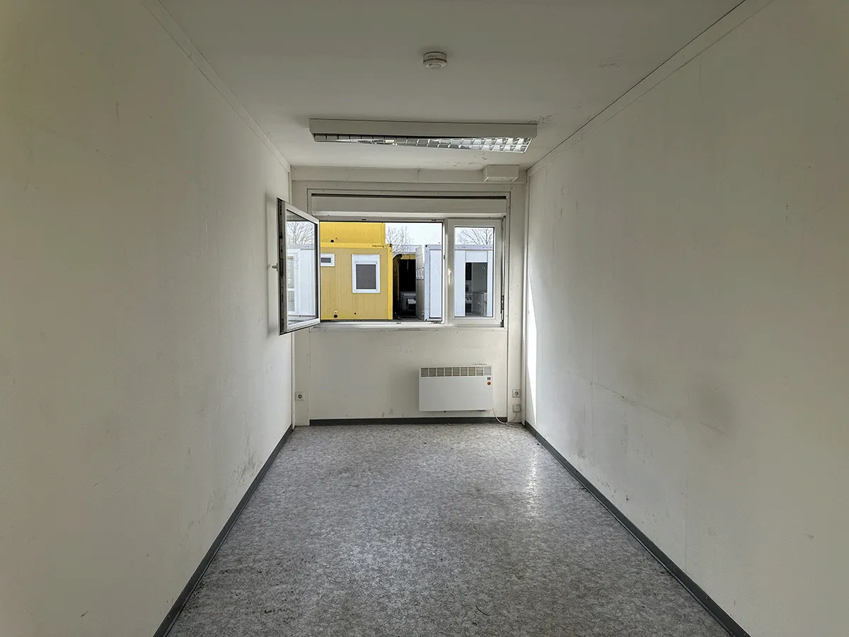 Innenansicht eines grauen Bürocontainers mit geöffnetem Fenster, Elektro-Heizkörper und hellem Bodenbelag.