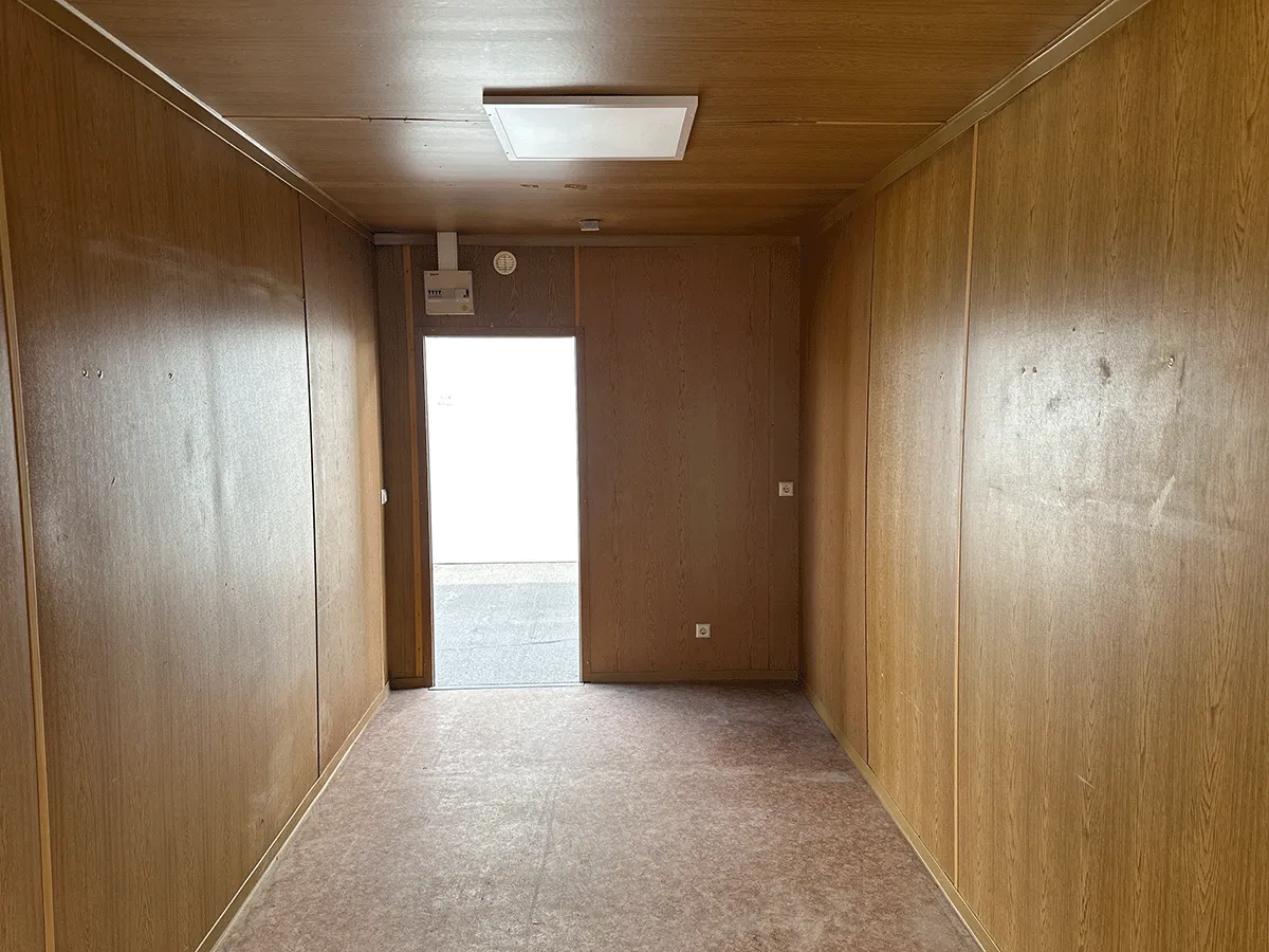 Innenansicht eines grauen Bürocontainers mit Fokus auf die geschlossene weiße Eingangstür und die Deckenbeleuchtung.