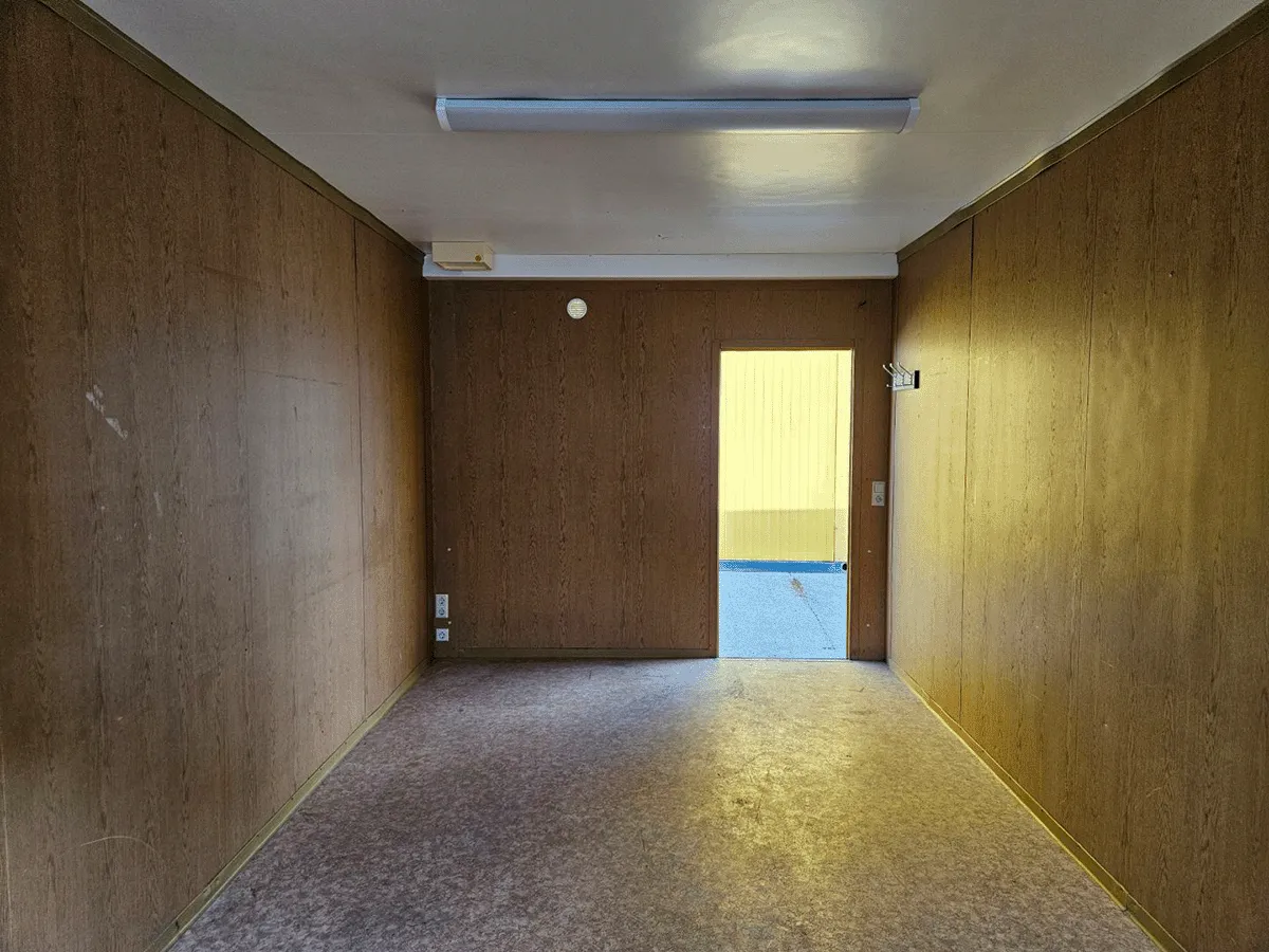 Innenansicht eines gelben Bürocontainers (3m Breite) mit Blick auf die geschlossene Eingangstür und die Deckenbeleuchtung.