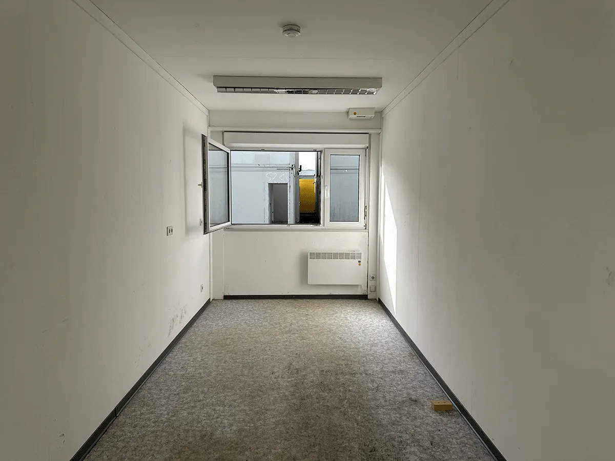 Innenansicht eines leeren grauen Bürocontainers mit Fenster, Heizkörper und hellem, gesprenkeltem Bodenbelag.