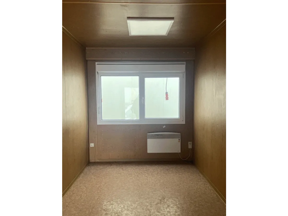 Innenansicht eines leeren grauen Bürocontainers mit Fenster, Elektro-Heizung und hellem Bodenbelag.