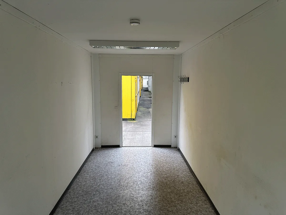 Innenansicht eines leeren grauen Bürocontainers mit Fenster, Heizkörper und hellem Bodenbelag.