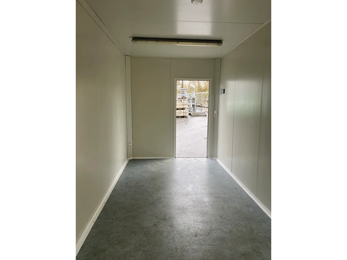 Innenansicht eines leeren, grauen Bürocontainers mit Blickrichtung zur Eingangstür und Deckenbeleuchtung.