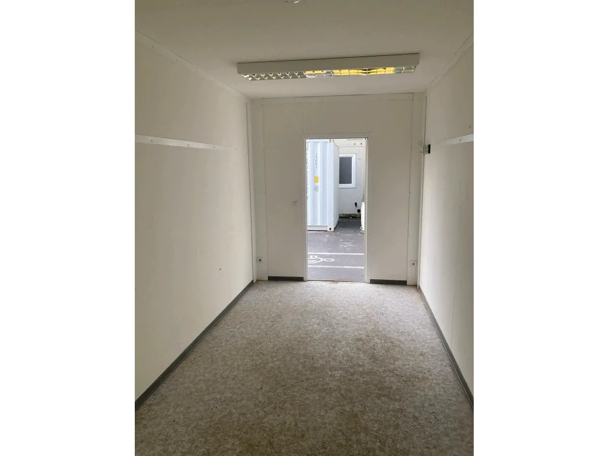 Innenansicht eines leeren grauen Bürocontainers mit Blick auf die geöffnete Tür und Deckenleuchte.